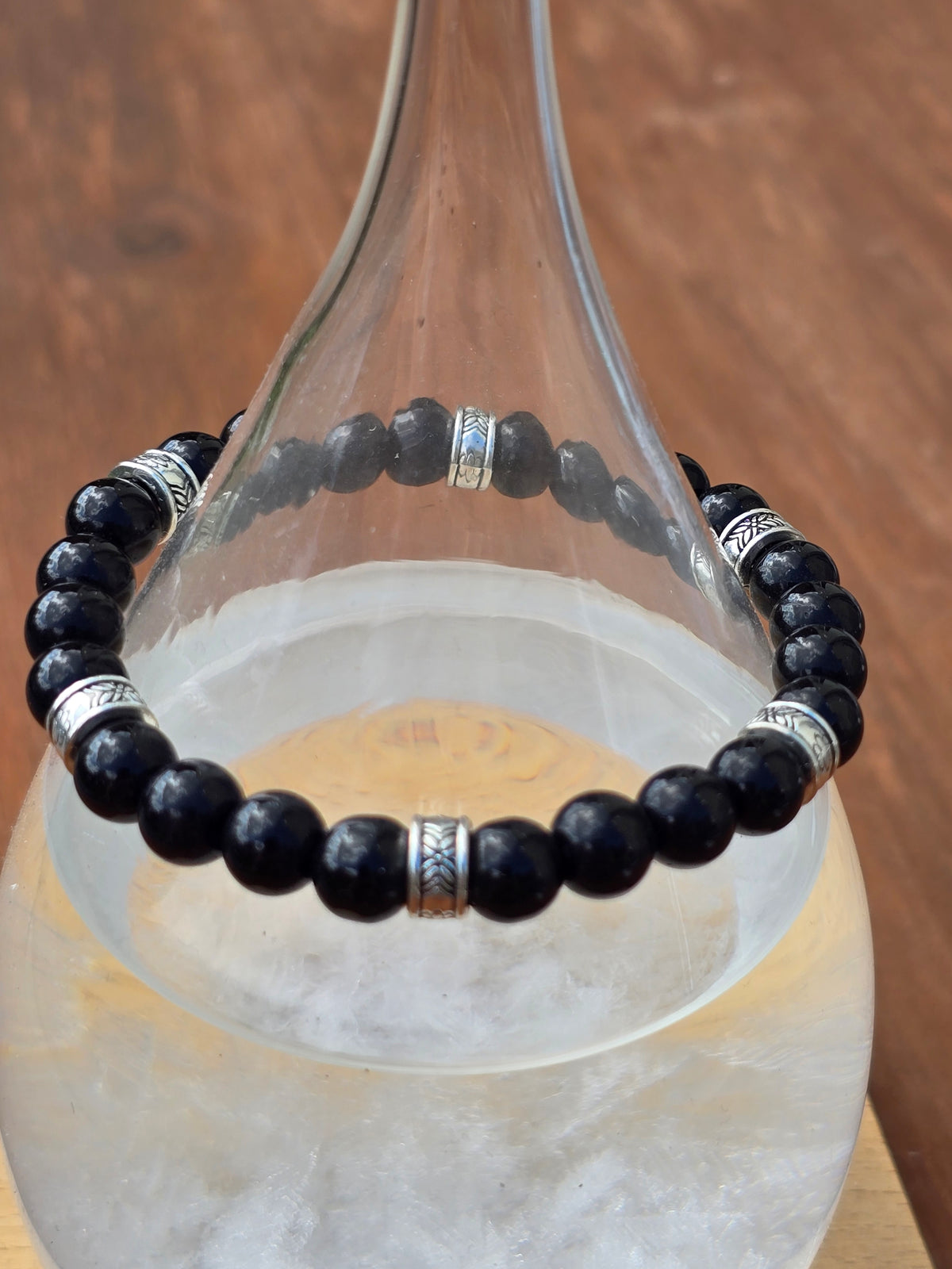 Glossy black beaded bracelet with engraved silver accents displayed around a clear glass decanter.