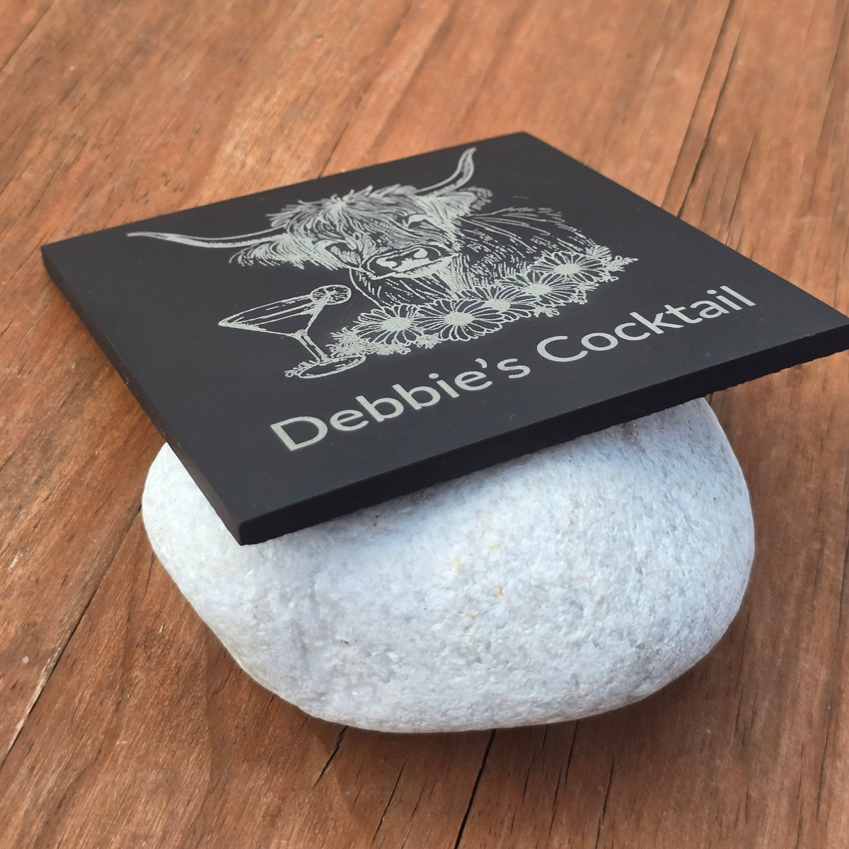 personalised slate coaster with highland cow and cocktail glass design sits against a white pebble stone