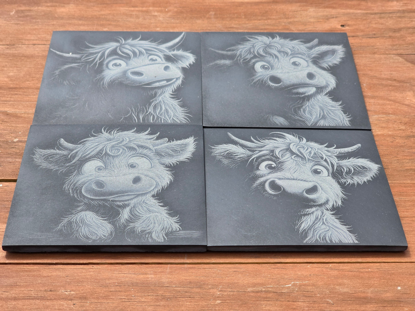 Four black slate coasters with highland cow illustrations on a wooden surface