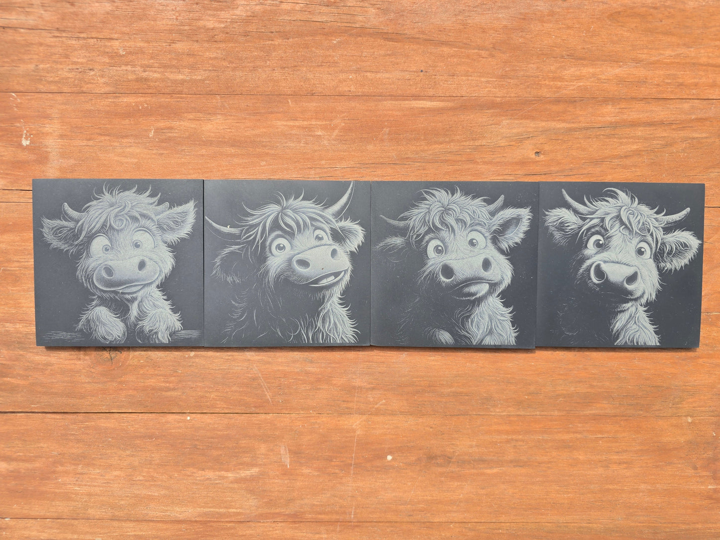 Four illustrations of a cartoon highland cow on black slate coasters, placed on a wooden surface.