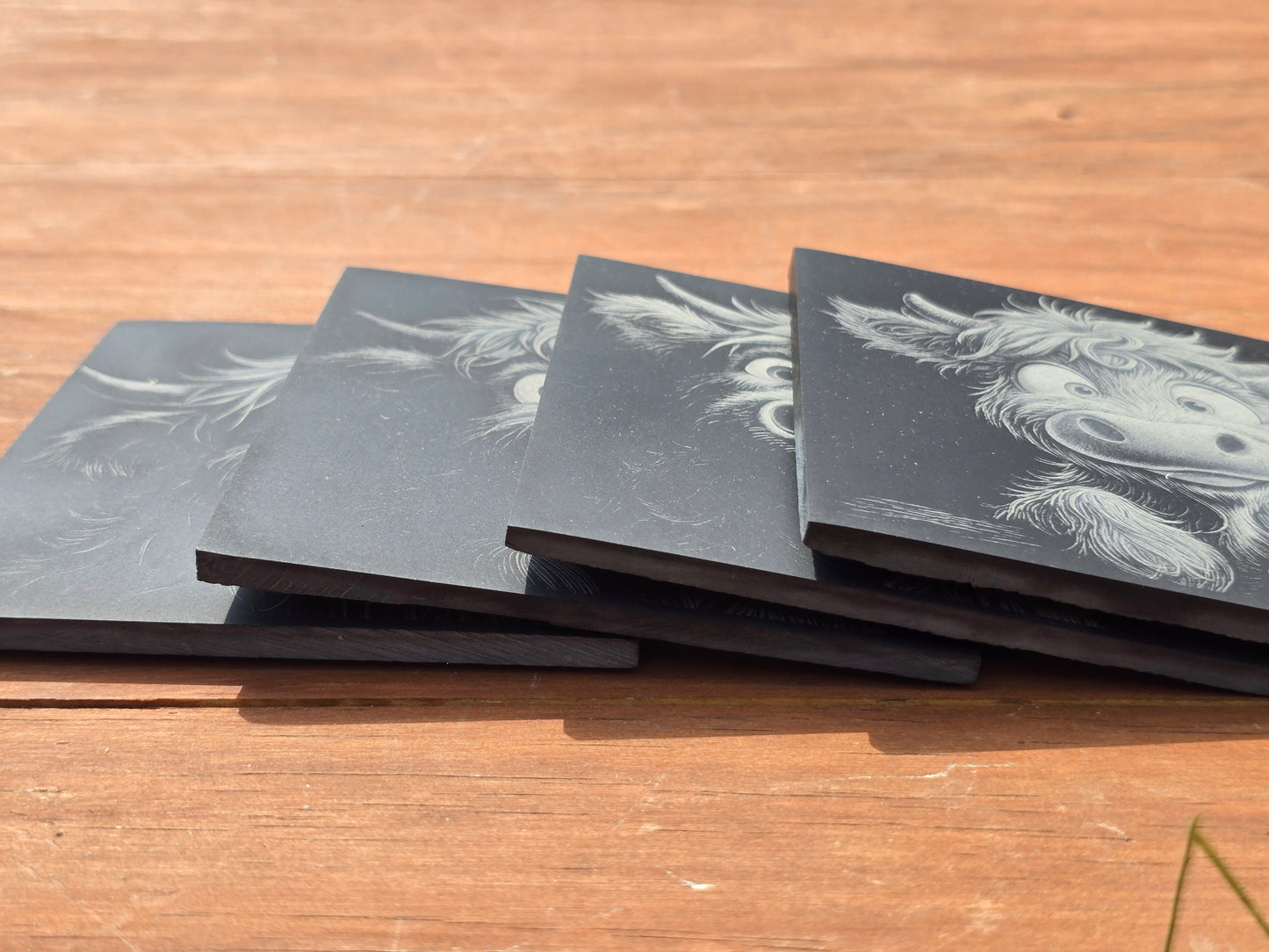 Four black coasters with highland cow designs on a wooden surface