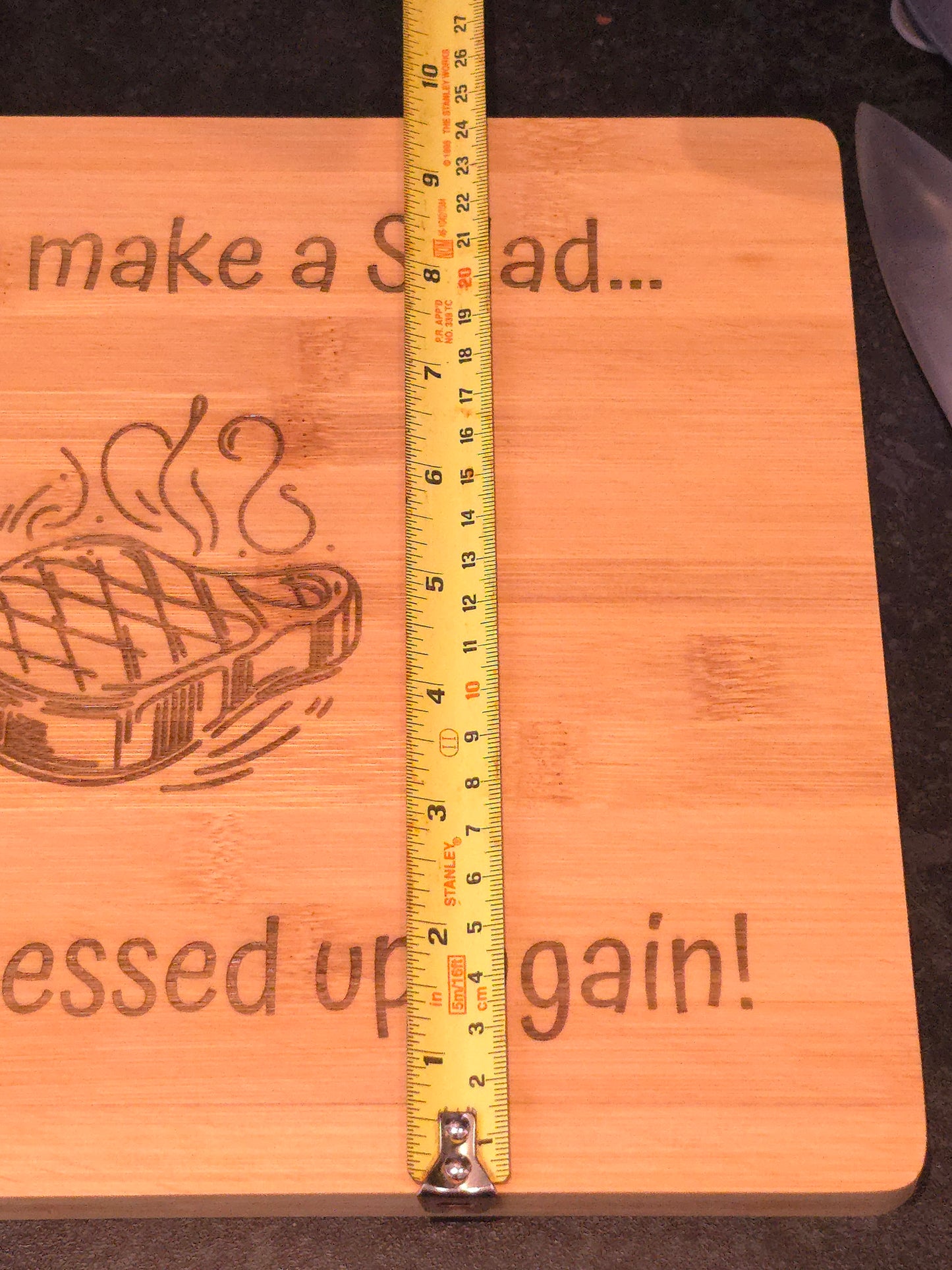 Wooden board with engraved text and design, measured with a tape measure, showing 24cm width