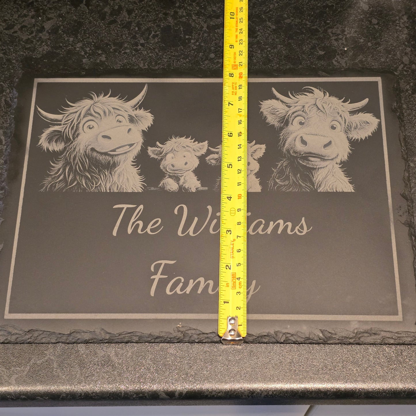 Decorative mat with engraved highland cows and 'The Williams Family' text, measured with a tape measure depicting 21cm width