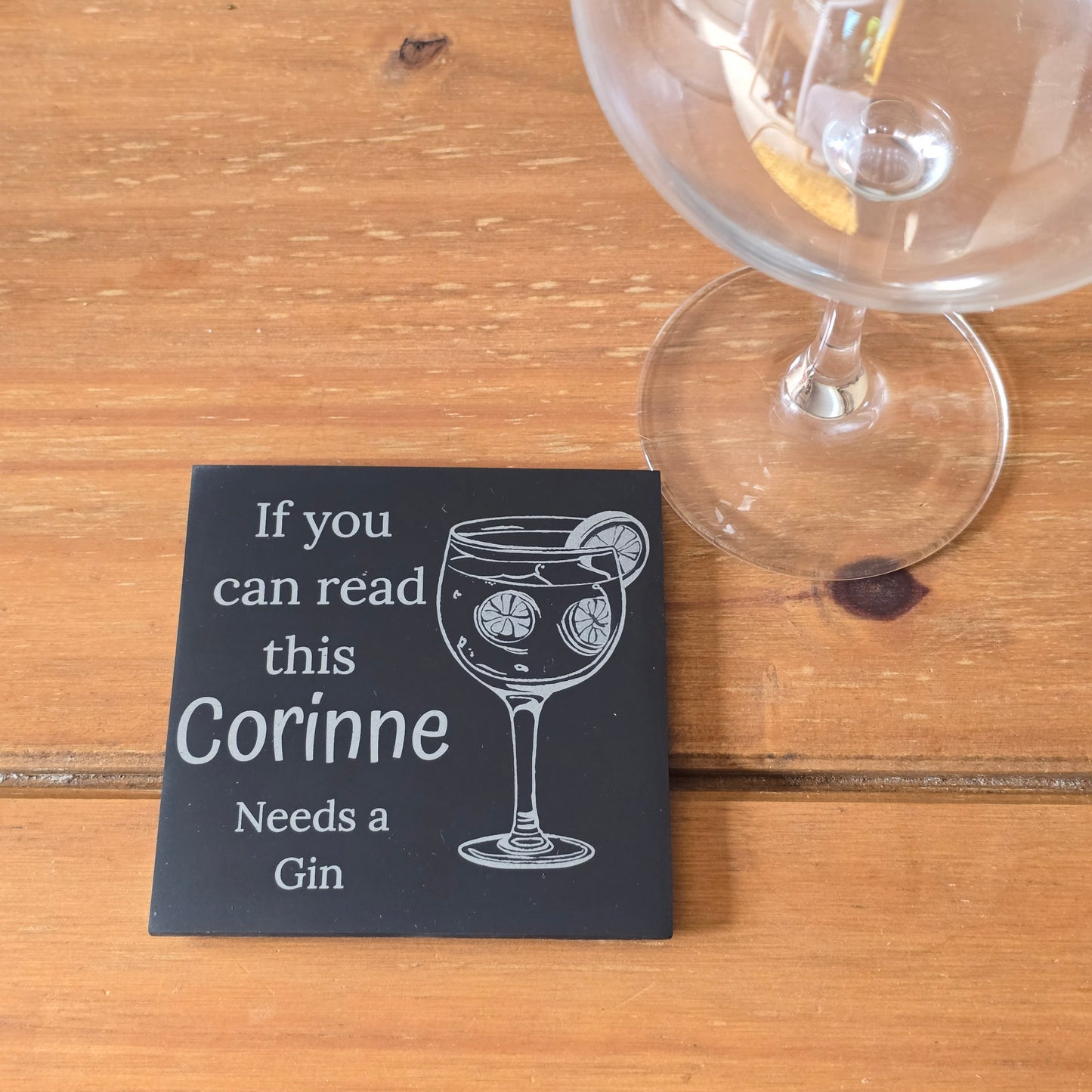 Slate coaster with text 'If you can read this Corinne Needs a Gin' next to a gin glass on a wooden surface