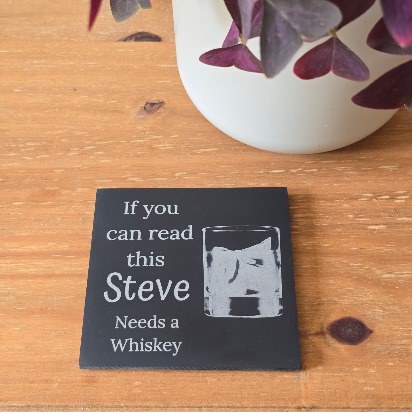 Black slate drinks mat with text 'If you can read this Steve needs a whiskey' on a wooden surface with a plant in the corner.