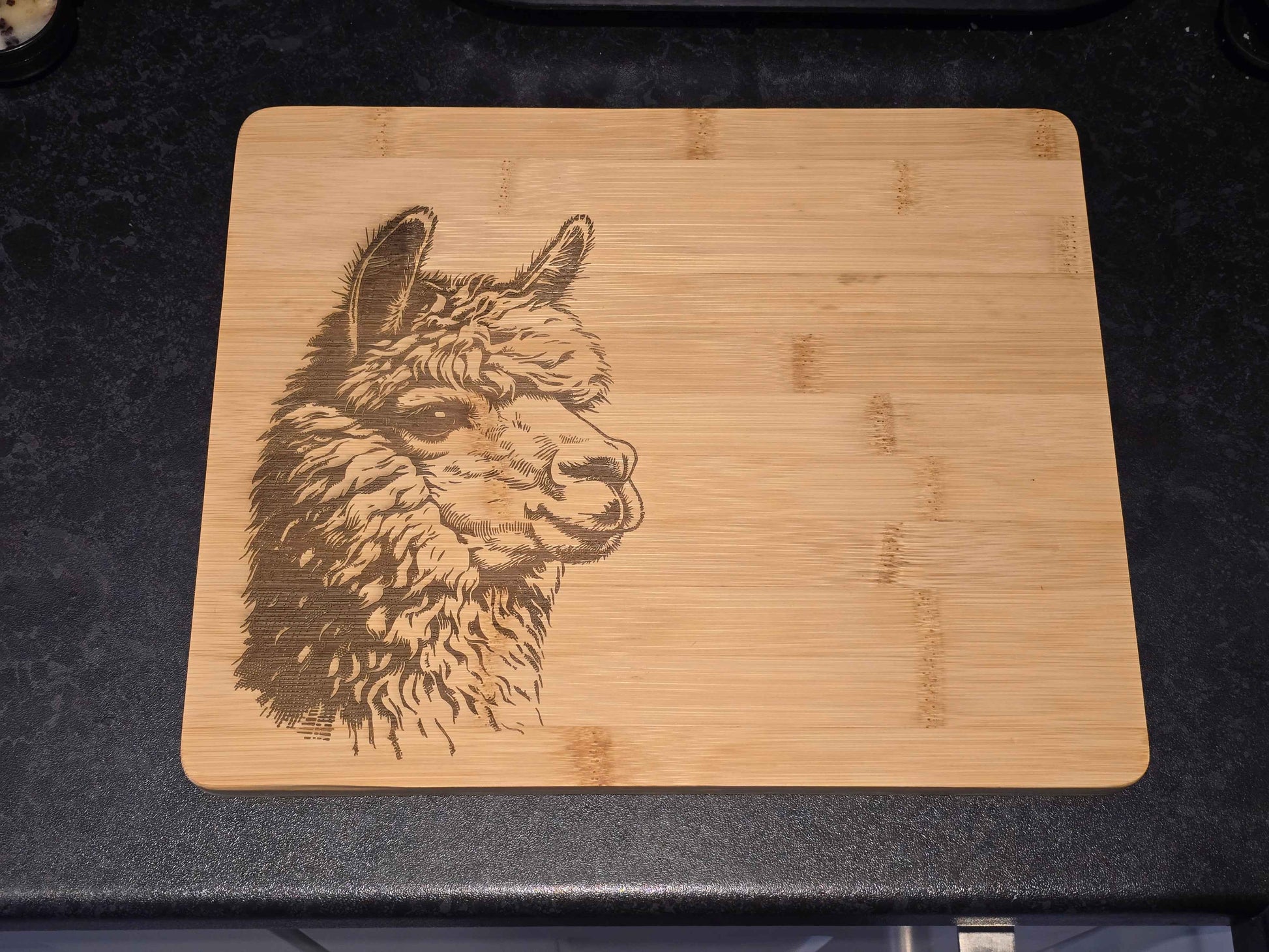 Bamboo cutting board with a laser-engraved alpaca design on a black countertop.
