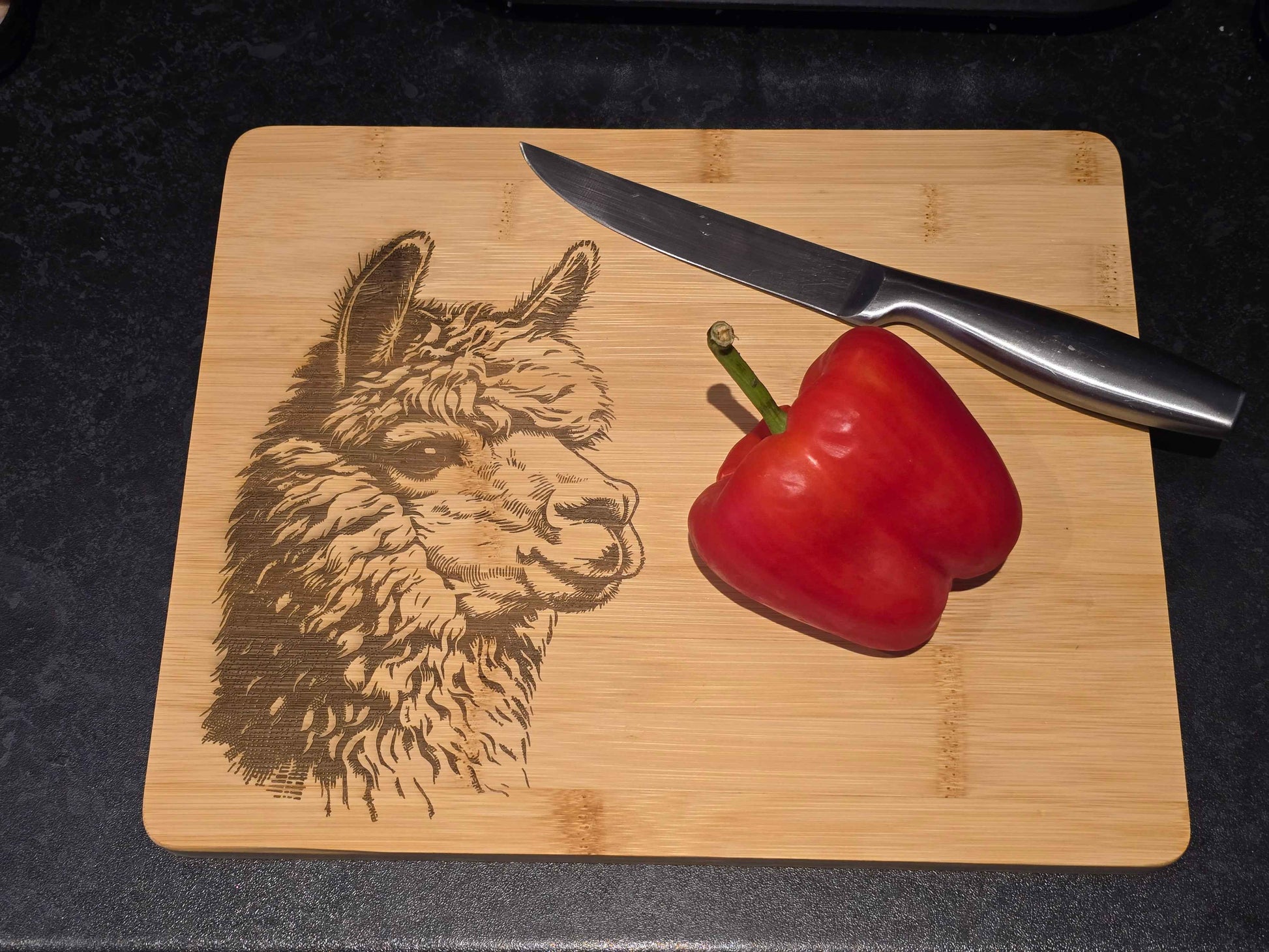 Bamboo cutting board with engraved alpaca design, knife, and red pepper on a dark surface