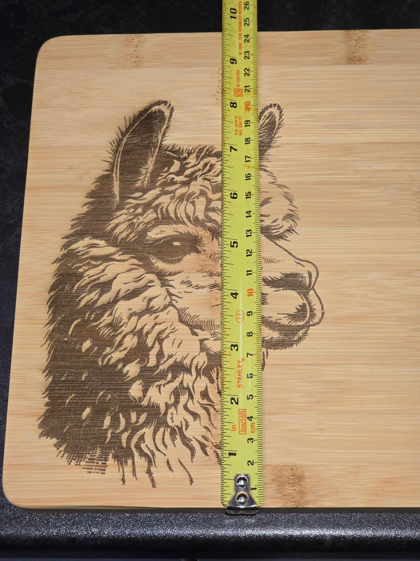 Bamboo cutting board with laser-engraved alpaca design and a tape measure for scale showing 24cm in width