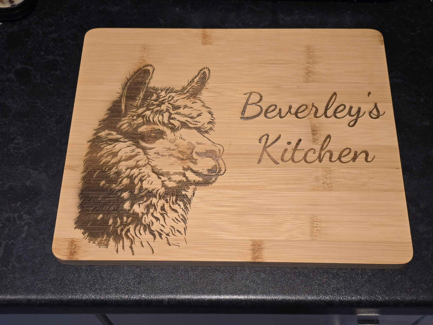 Personalised Bamboo cutting board with engraved alpaca design and 'Beverley's Kitchen' text on a dark surface.