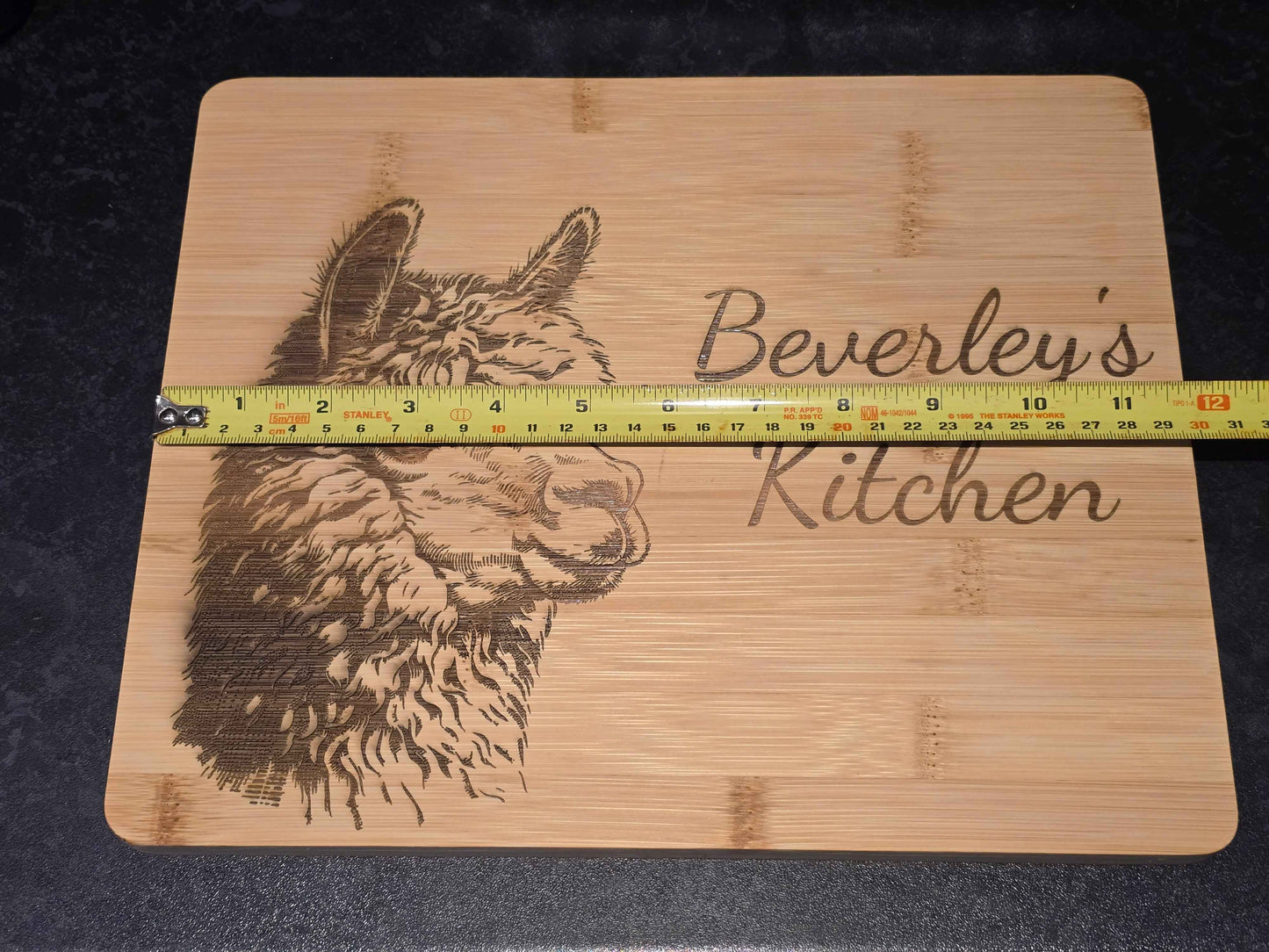 Bamboo cutting board with engraved alpaca design and 'Beverley's Kitchen' text, measured by a ruler showing 30cm length