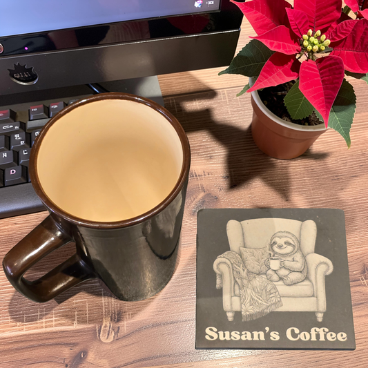 A dark brown ceramic coffee mug sits on a wooden desk next to an engraved slate coaster that reads 'Susan's Coffee' and shows a sloth in an armchair.