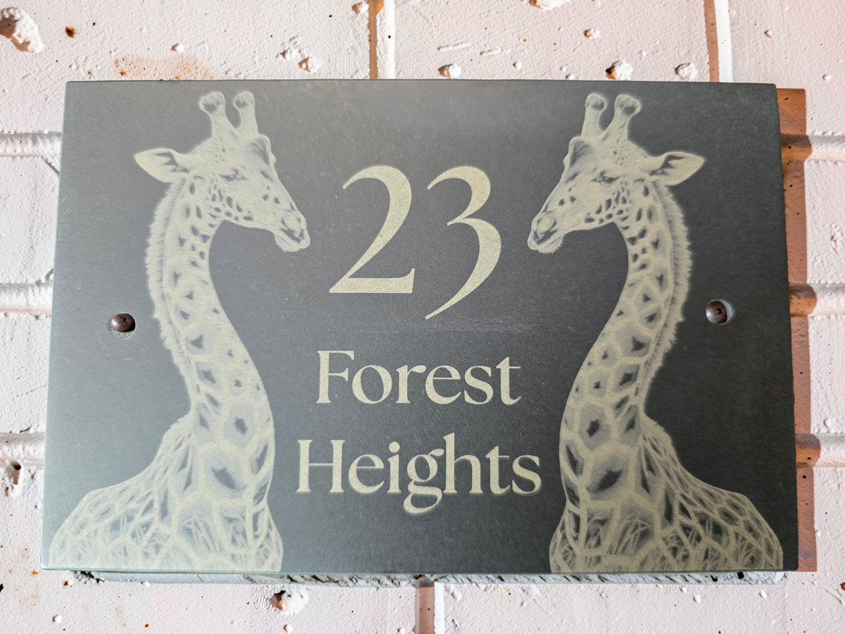 A rectangular dark gray house number sign featuring two stylized giraffe illustrations and the text '23 Forest Heights' in light beige, mounted on a white brick wall.