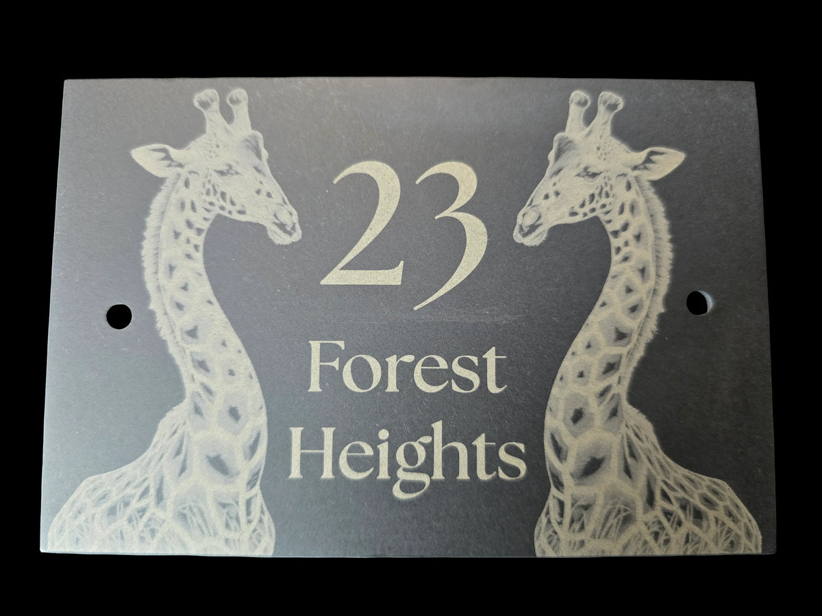 A rectangular dark gray plaque or sign featuring two engraved white giraffe illustrations and the text '23 Forest Heights' in white, shown against a black background.