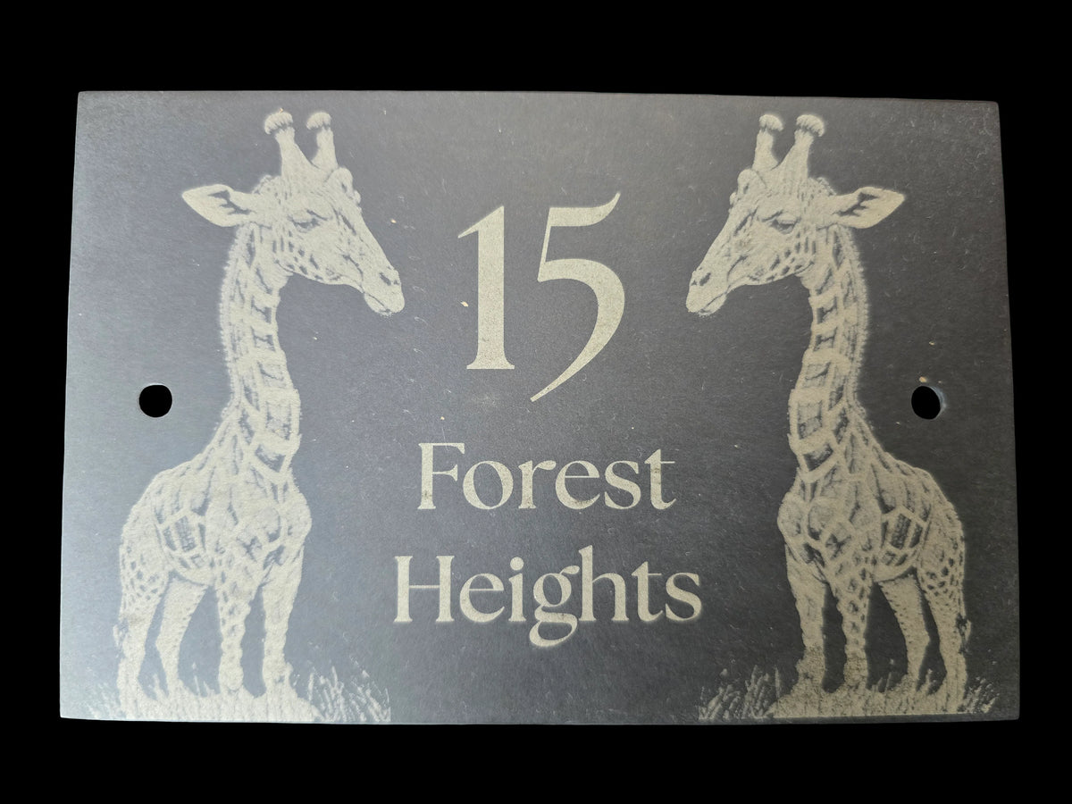 A rectangular dark grey house plaque or sign featuring two white giraffe illustrations and the text '15 Forest Heights' in white, shown against a black background.