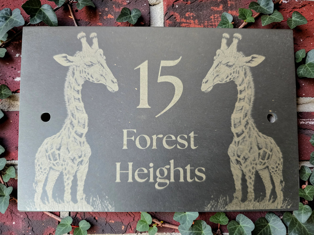 A rectangular dark slate house number plaque featuring two giraffe illustrations and the text '15 Forest Heights' mounted on a red brick wall with green ivy leaves around it.