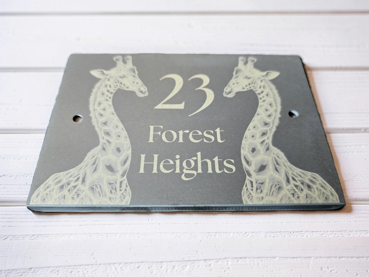 A custom rectangular dark slate house number plaque featuring two giraffe illustrations and the text '23 Forest Heights' in a light cream color, displayed on a white wood plank surface.