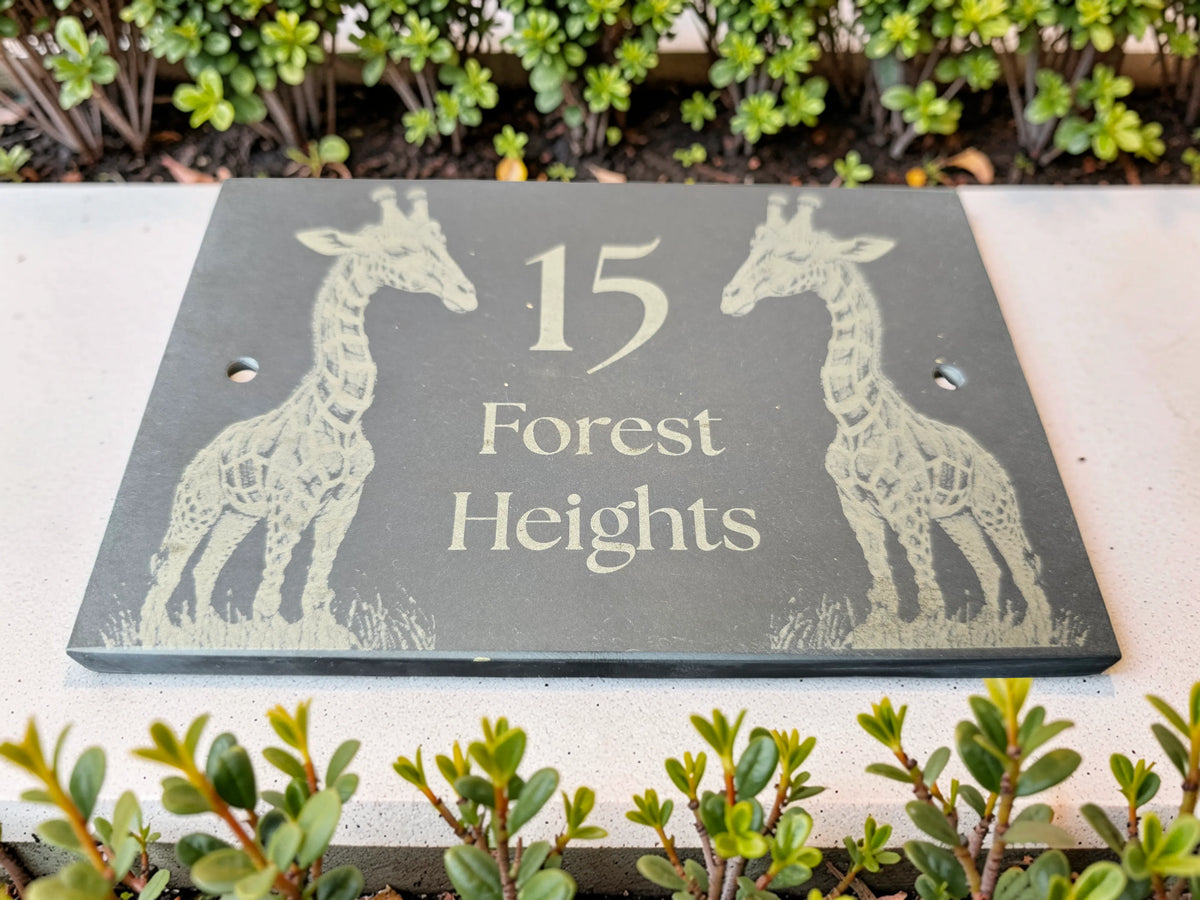 A rectangular dark slate memorial or garden plaque with engraved light-colored giraffe illustrations and the text '15 Forest Heights,' photographed outdoors among green shrubs.