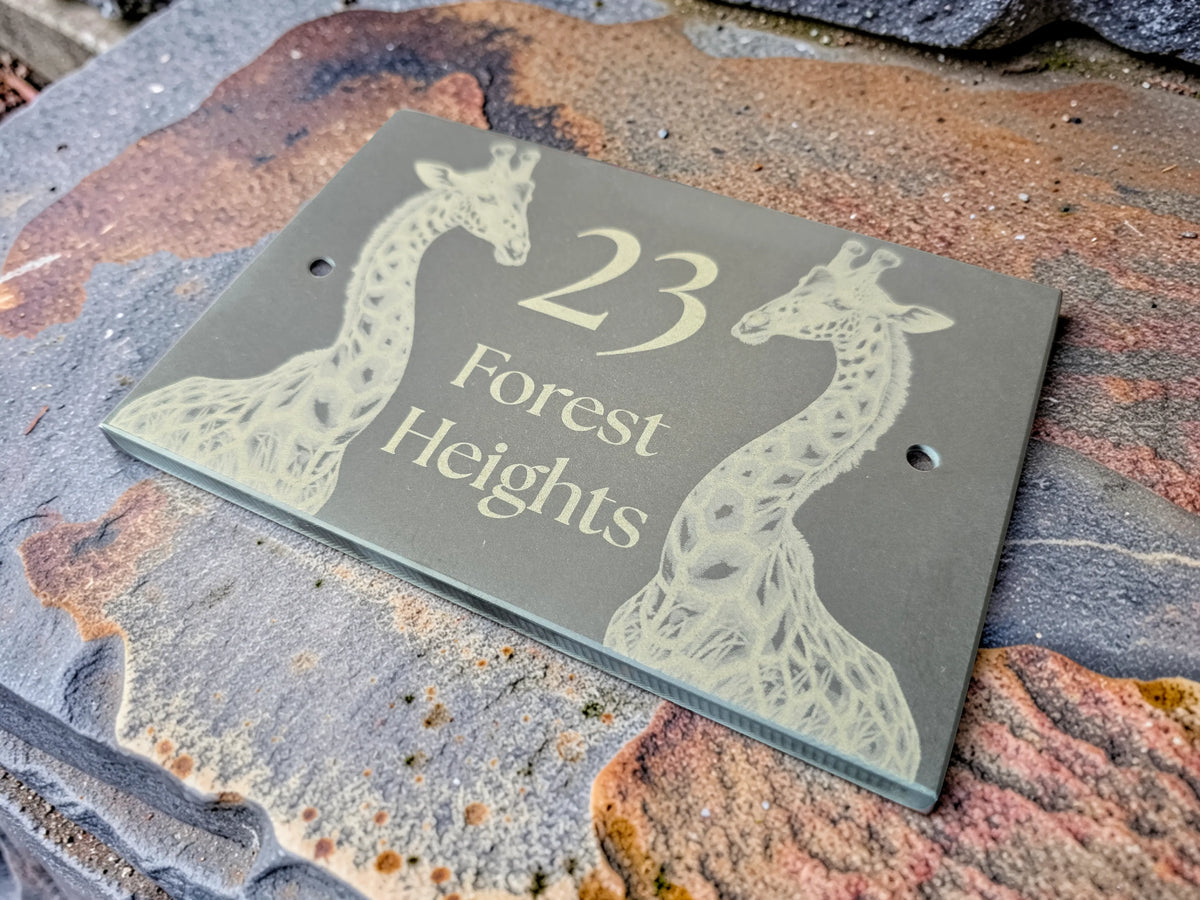 A personalised rectangular slate house number plaque with two engraved giraffe illustrations and the text '23 Forest Heights,' shown resting on a textured stone surface.