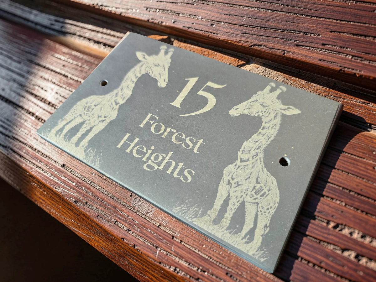 A rectangular dark grey slate house number plaque with engraved light beige giraffe illustrations and the text '15 Forest Heights,' shown mounted on a wooden surface in warm sunlight.