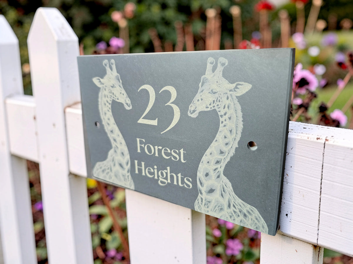 A rectangular dark gray house number plaque with two white giraffe silhouettes and the text '23 Forest Heights,' mounted on a white picket fence with a blurred garden background.