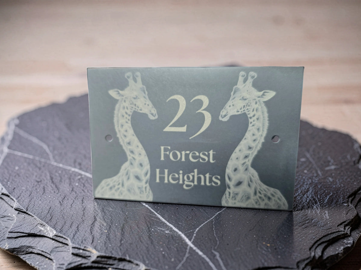 A rectangular slate house number sign with two giraffe engravings and the text '23 Forest Heights,' displayed on a dark slate coaster against a light wood background.