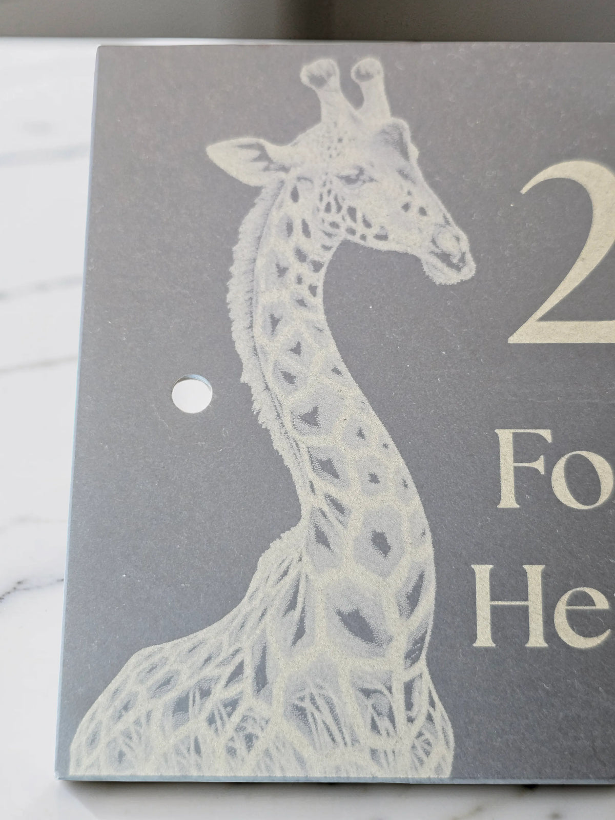 A close-up of a dark grey square sign or plaque featuring a white giraffe illustration placed on a light marble-like surface.