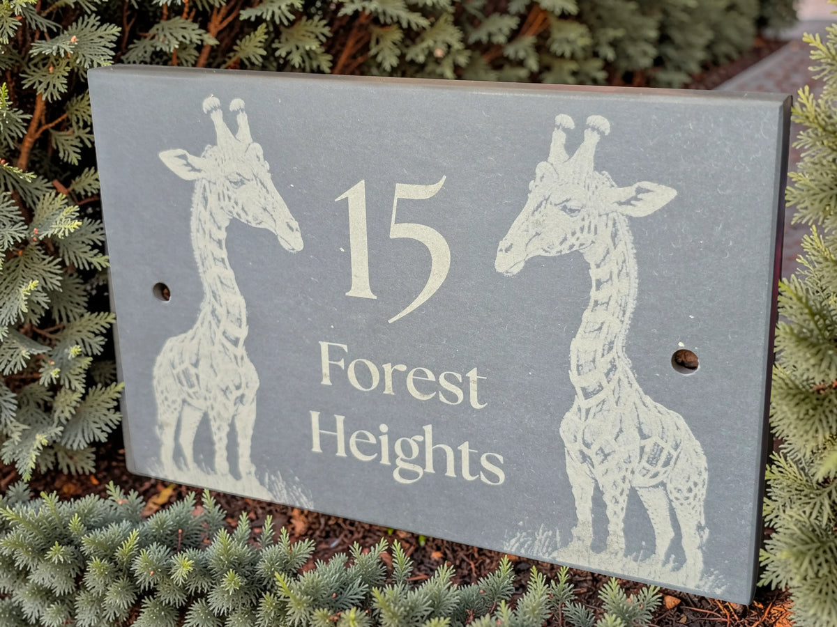 A rectangular grey slate plaque or sign with two white giraffe illustrations and the text '15 Forest Heights,' photographed outdoors among green plants.