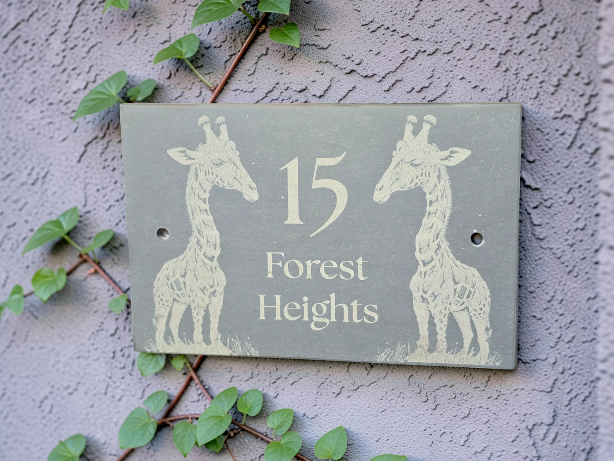 A personalised rectangular grey house number plaque mounted on a textured stucco wall, featuring two cream giraffe silhouettes and the text '15 Forest Heights.' Green ivy vines are visible around the plaque.