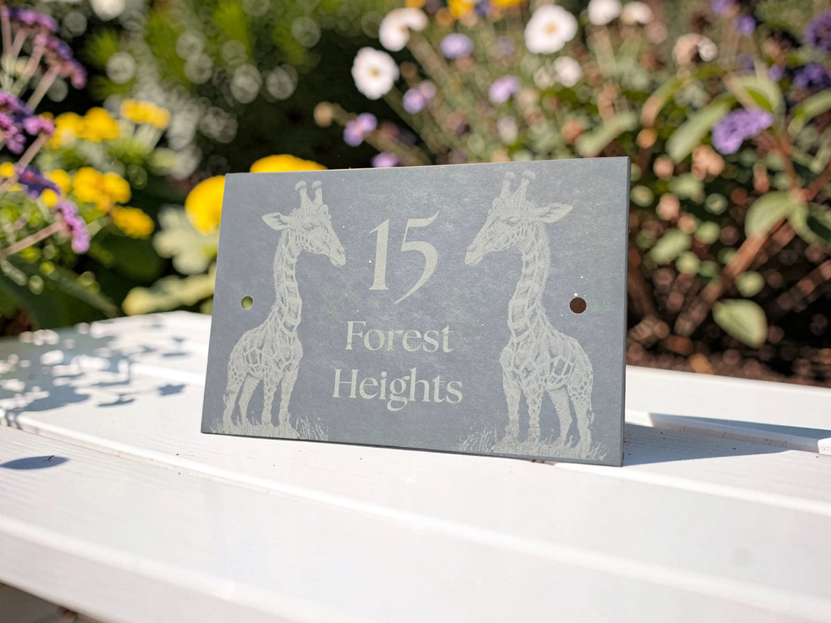 A rectangular slate house number sign with engraved giraffe illustrations and the text '15 Forest Heights', displayed on a white wooden fence with a blurred garden background.