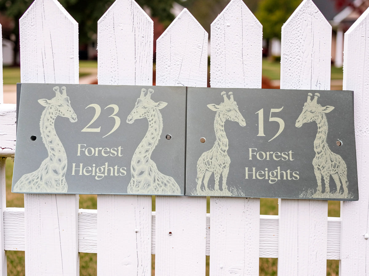 Two rectangular dark grey slate house numbers for '23 Forest Heights' and '15 Forest Heights' with giraffe illustrations, mounted on a white picket fence.