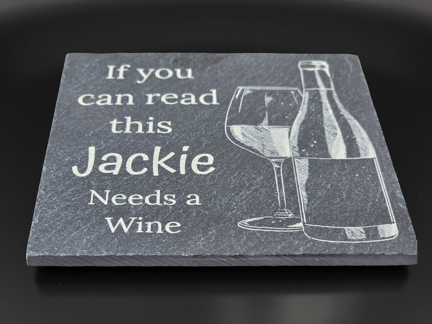 A personalized slate coaster with the text 'If you can read this Jackie Needs a Wine' engraved, along with an image of a wine bottle and glass.