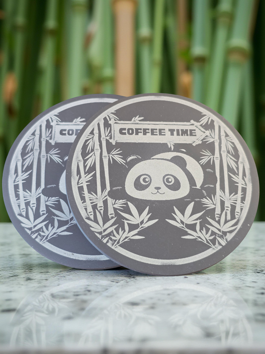 Two grey slate coasters with panda and bamboo design on a marble surface with a bamboo background.