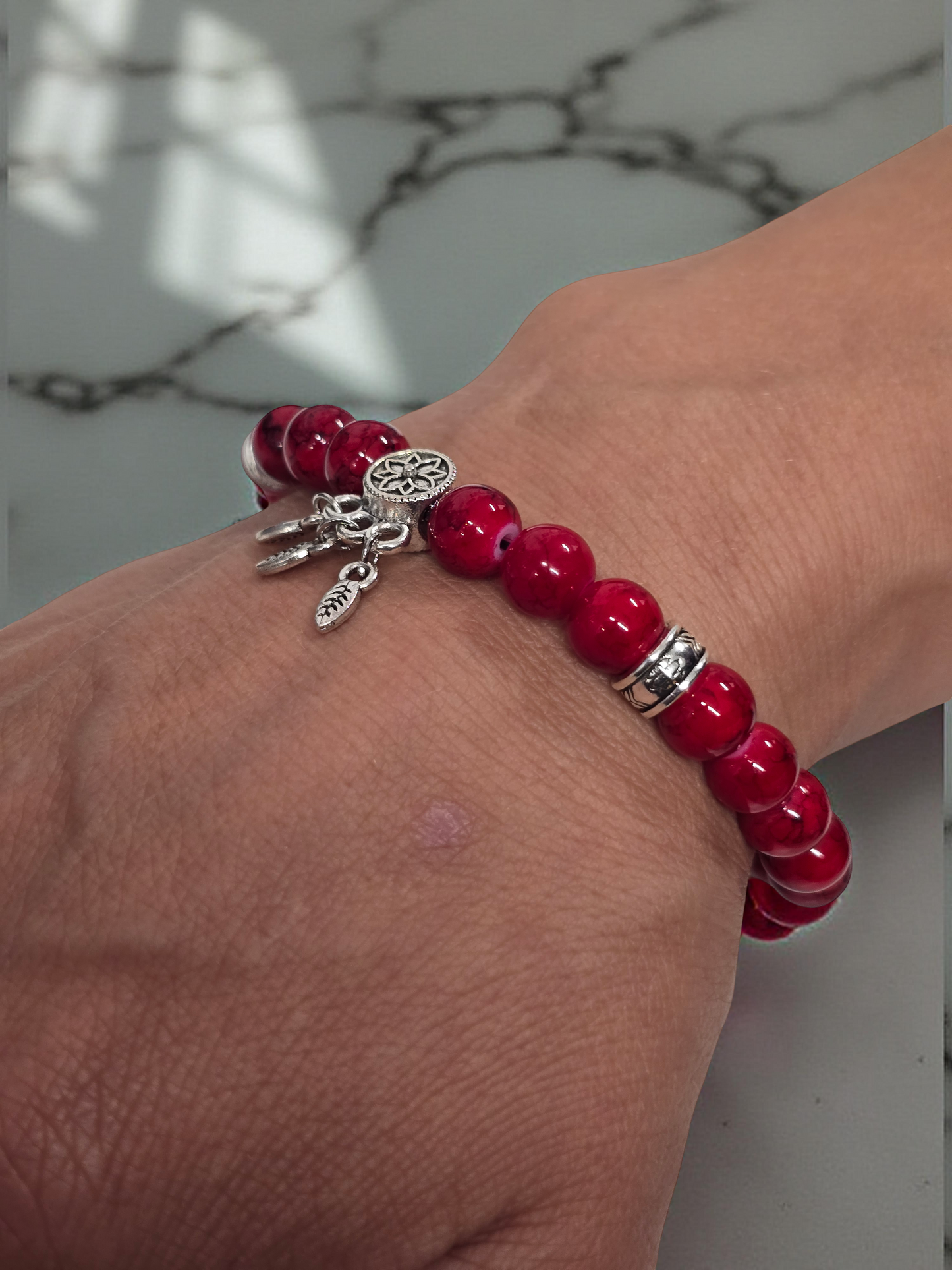 Red beaded bracelet with silver charms and dreamcatcher dangle charm on a wrist against a light marble background