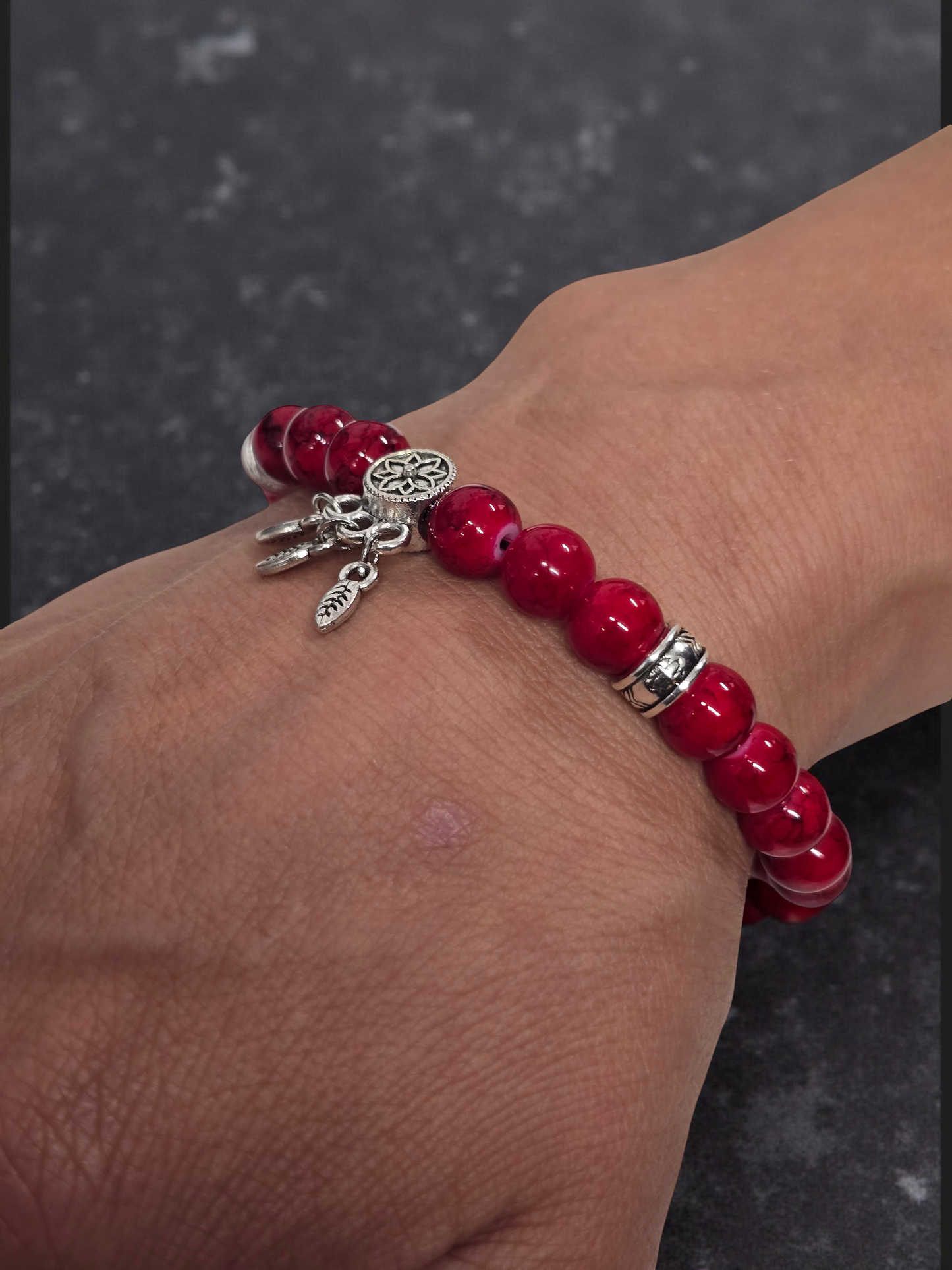 Red beaded bracelet with silver charms shown on a wrist against a dark marble background