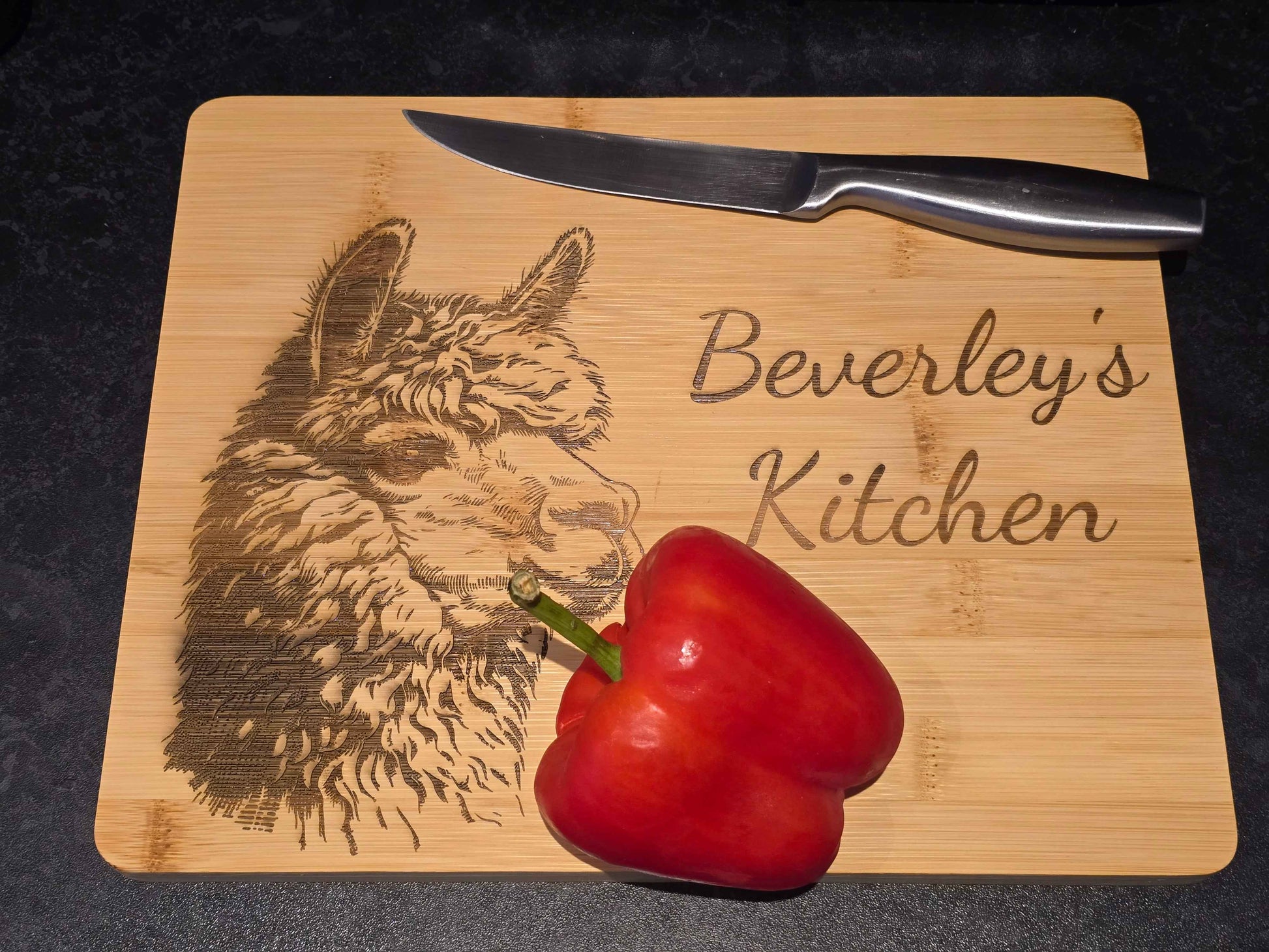 Bamboo cutting board with engraved alpaca design and 'Beverley's Kitchen' text, accompanied by a red pepper and knife.