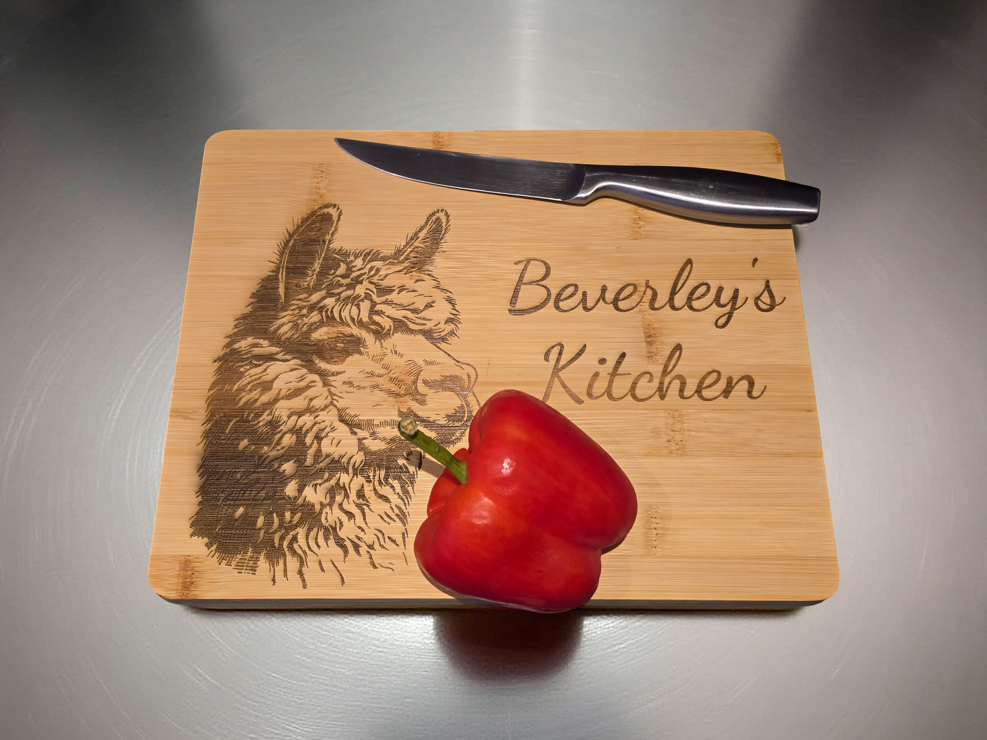 Customised Bamboo cutting board with engraved alpaca design and 'Beverley's Kitchen' text, knife, and red pepper on a metallic surface.