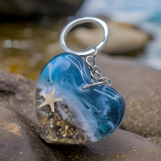 A heart-shaped resin keyring with a starfish, featuring real sand and shells, placed on a rocky surface with a blurred background.