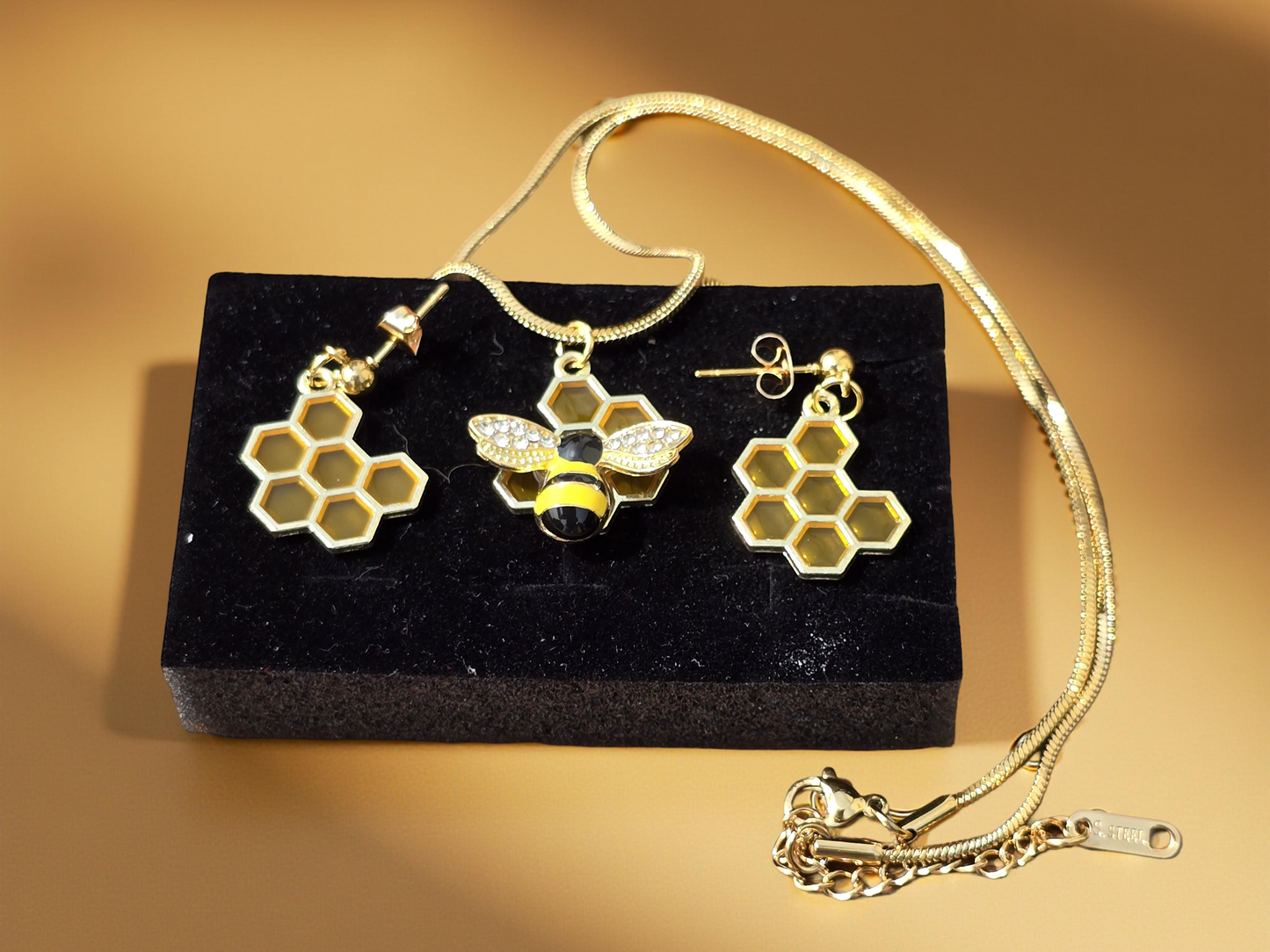 A gold-colored necklace with a bumblebee and honeycomb pendant, accompanied by a pair of honeycomb earrings, displayed on a black cushion.