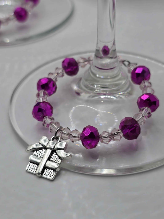 Silver gift box charm on a purple and clear beaded ring wrapped around a wine glass stem.