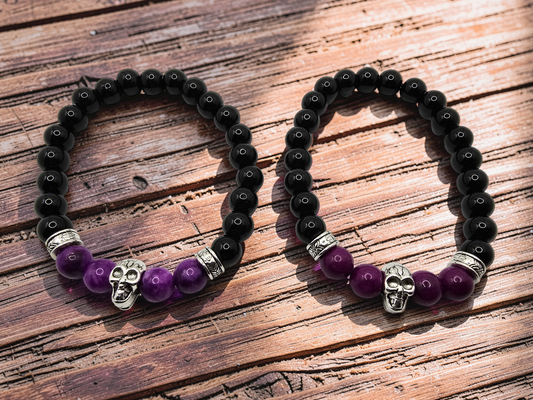Two beaded stretch bracelets with black round beads, purple amethyst beads, silver spacer rings, and skull charms, displayed on a rustic wooden surface.