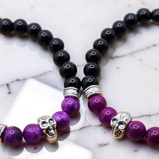 A close-up of a beaded bracelet laid on a white marble surface, featuring glossy black round beads, purple stone beads, silver spacer rings, and small metallic skull charms.