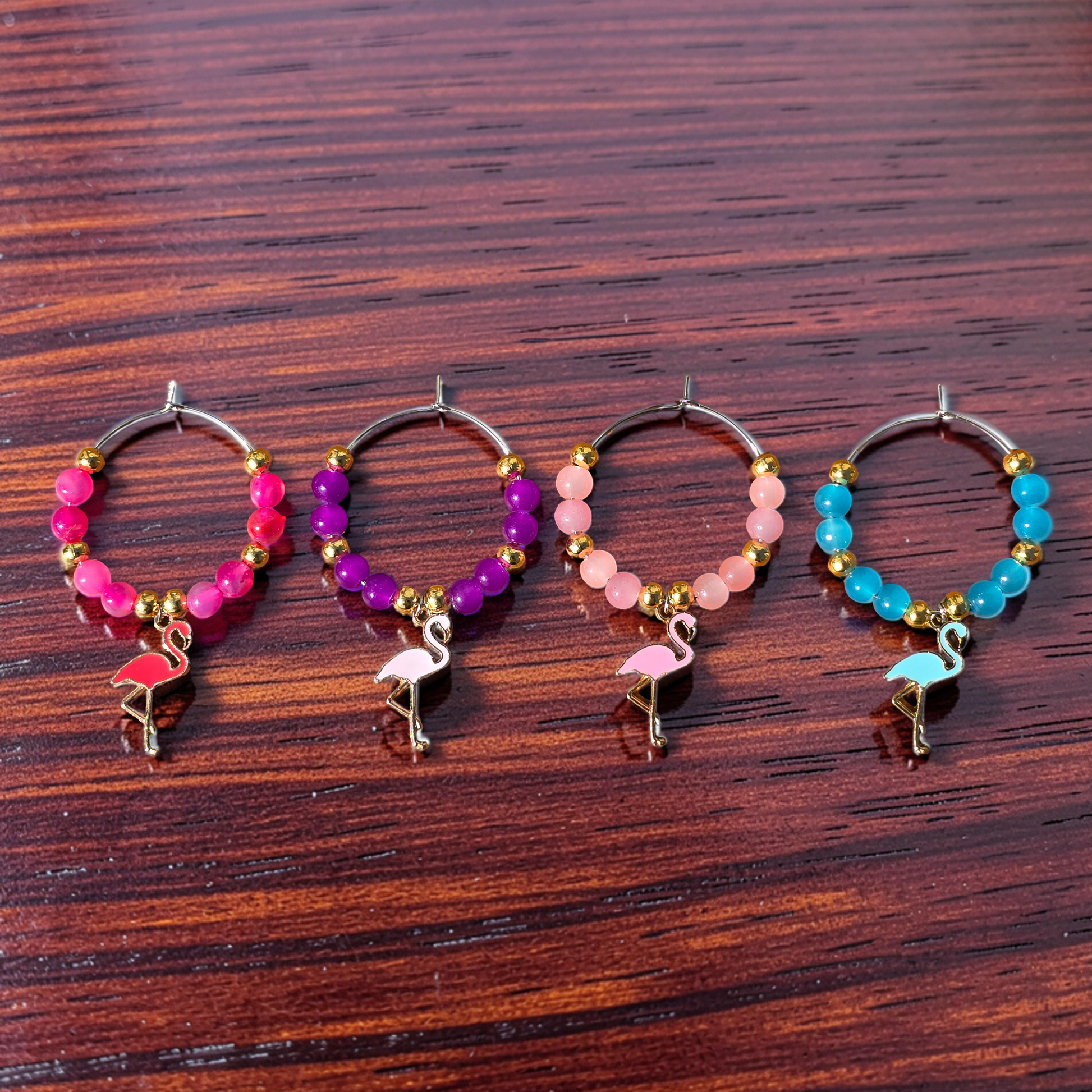 Four beaded wine glass charms with flamingo accessories, displayed on a dark wood surface. The hoops feature different colour bead combinations: pink, purple, light pink, and blue, with gold-toned metal and small flamingo charms.