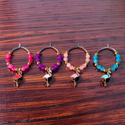 Four beaded wine glass charms with flamingo accessories, displayed on a dark wood surface. The hoops feature different colour bead combinations: pink, purple, light pink, and blue, with gold-toned metal and small flamingo charms.