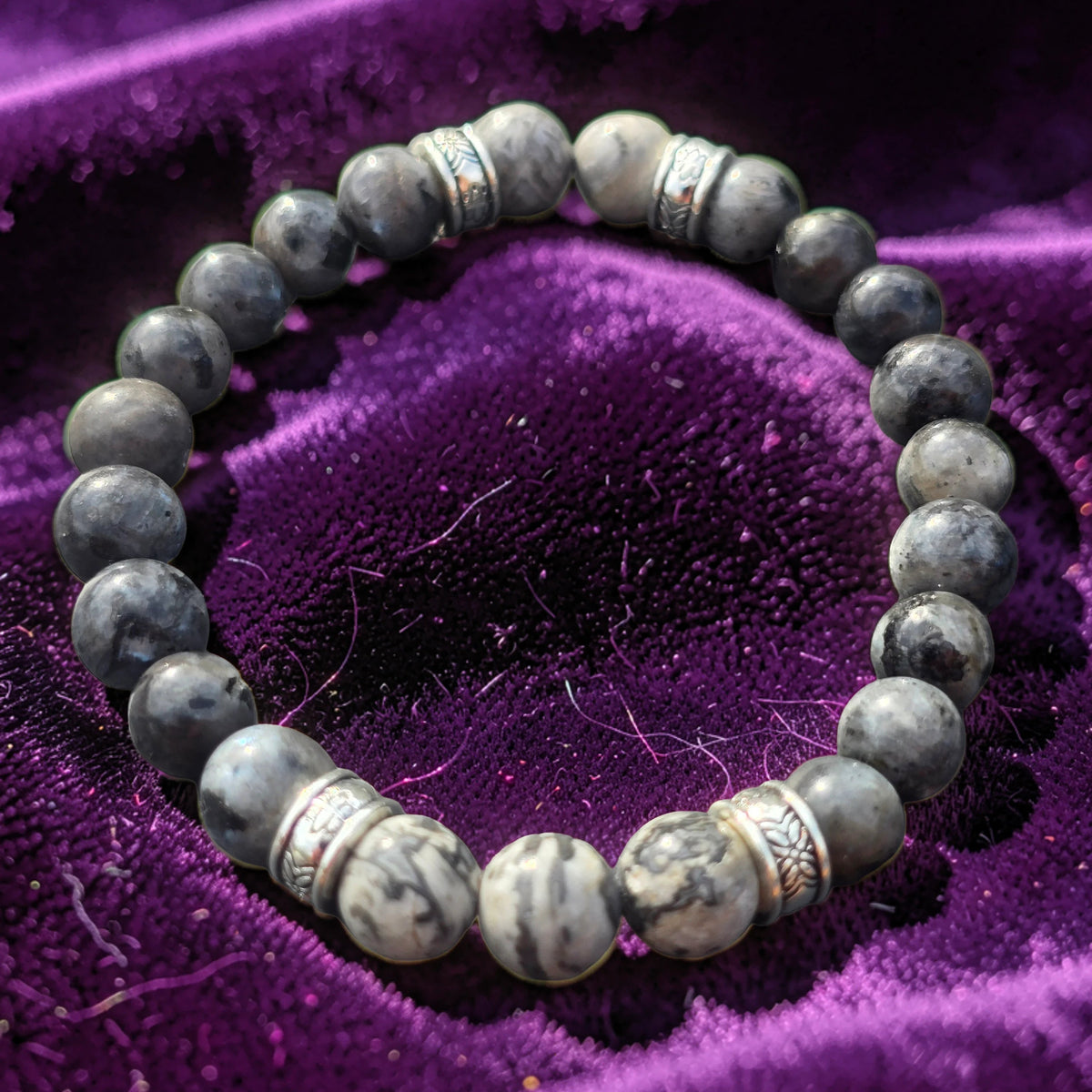 A beaded stretch bracelet made of polished gray stone beads with silver-toned spacer accents, displayed on a textured purple velvet surface.