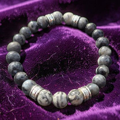 A beaded stretch bracelet made of polished gray stone beads with silver-toned spacer accents, displayed on a textured purple velvet surface.