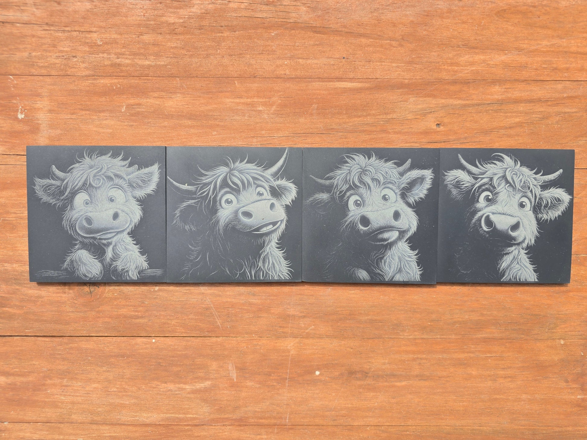 Four illustrations of a cartoon highland cow on black slate coasters, placed on a wooden surface.