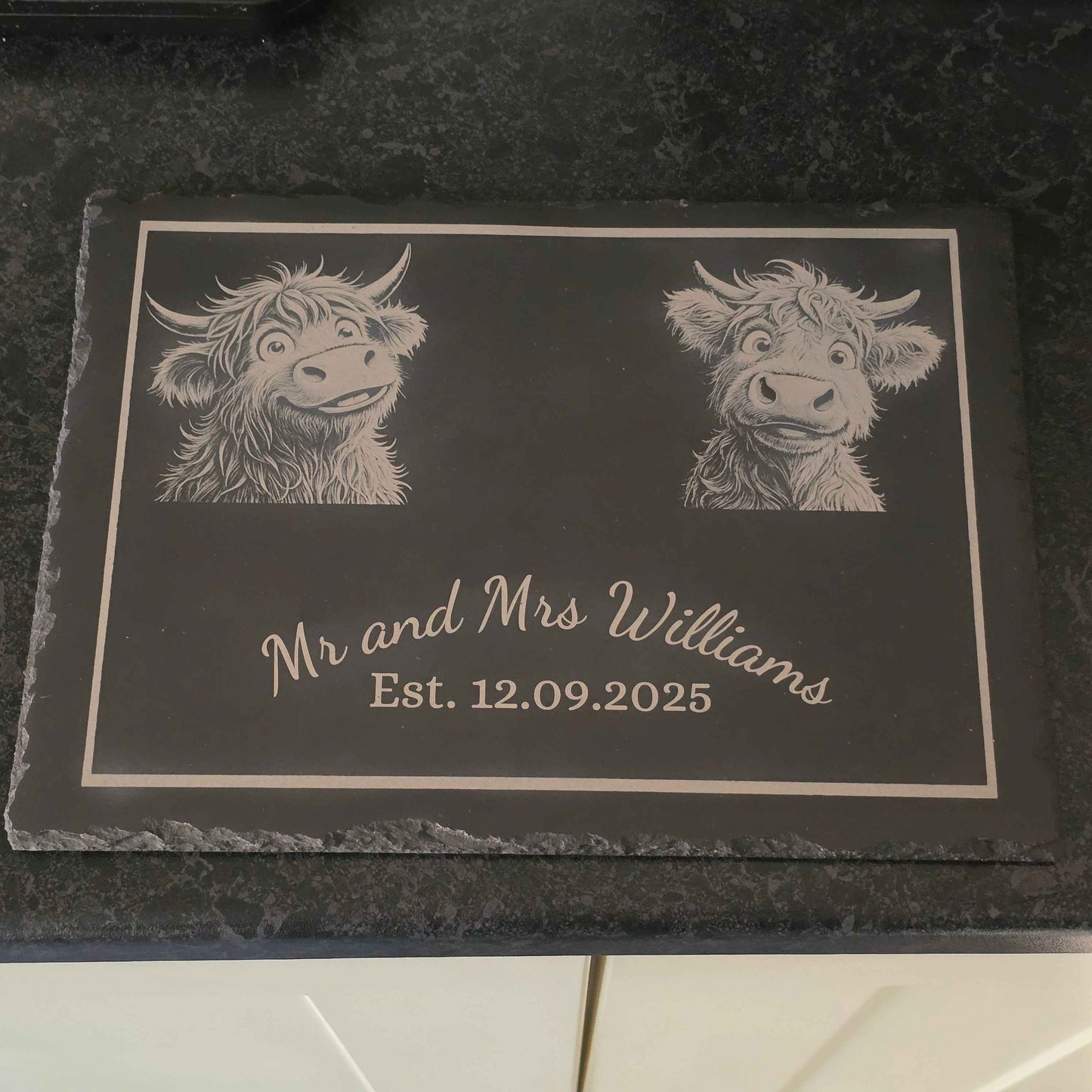 Personalized slate plaque with engraved highland cow illustrations and text on a dark surface.