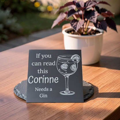 custom slate coaster with text and a gin glass engraving on a wooden table with a plant in the background.