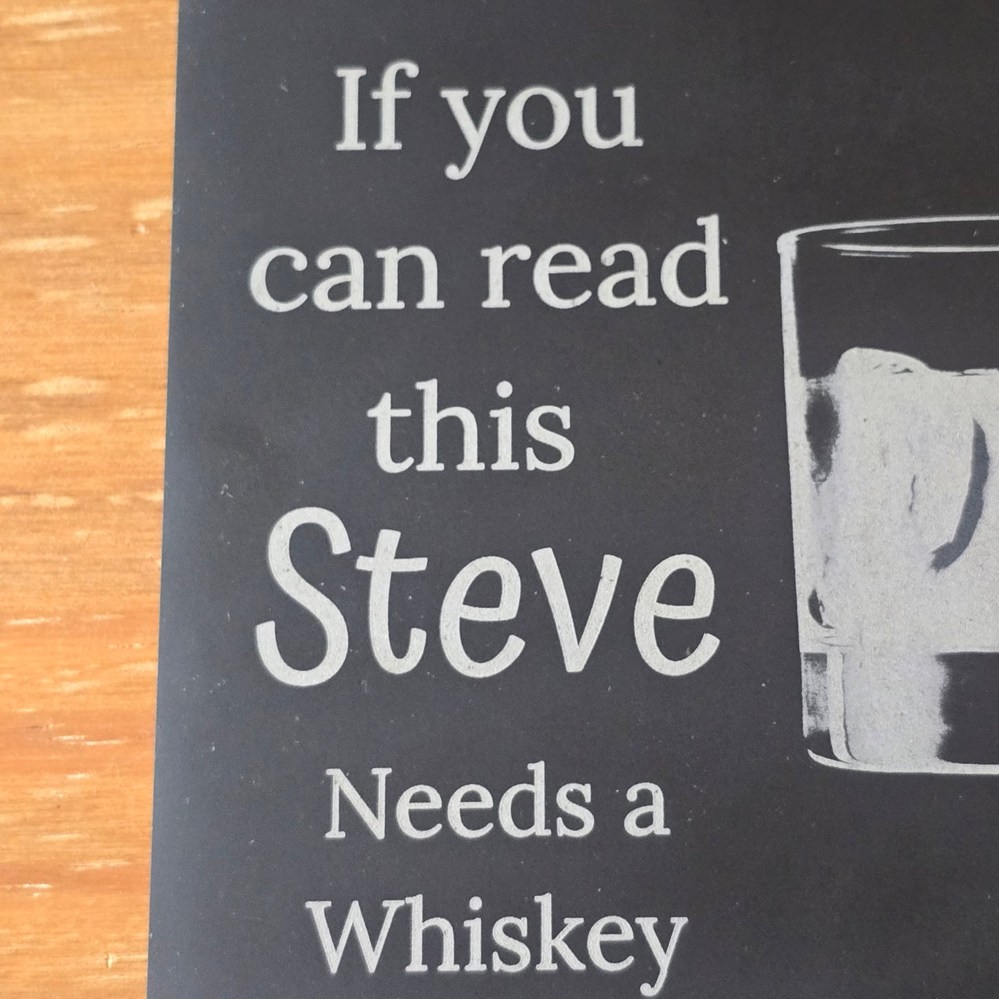 Black coaster with white text 'If you can read this Steve Needs a Whiskey' on a wooden surface.