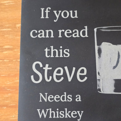 Black coaster with white text 'If you can read this Steve Needs a Whiskey' on a wooden surface.